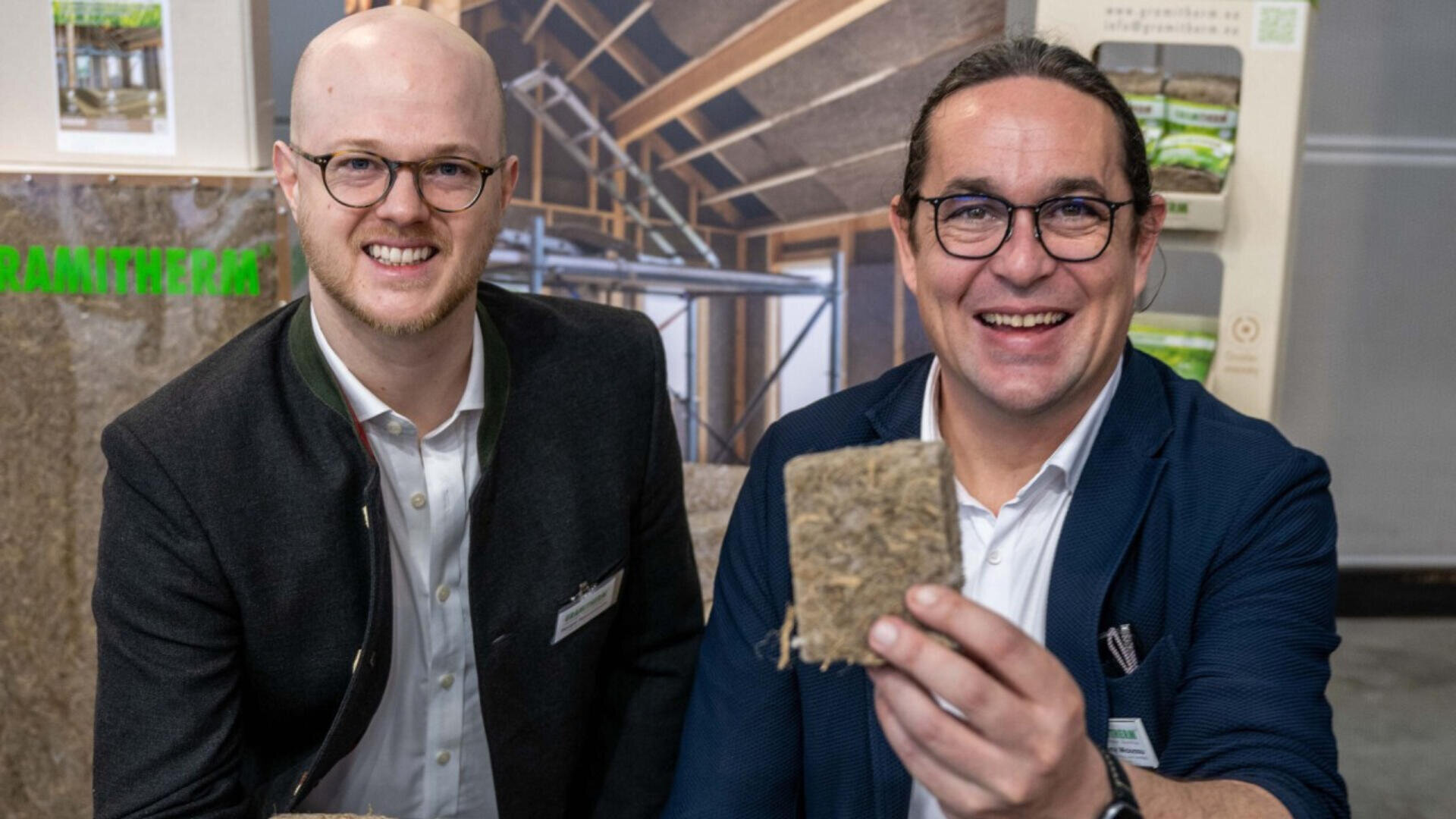 Zwei lächelnde Männer in Geschäftskleidung halten an einem Messe- oder Ausstellungsstand ein quadratisches Stück faseriges Isoliermaterial hoch, im Hintergrund sind Baumaterialien und eine Anzeigetafel zu sehen.