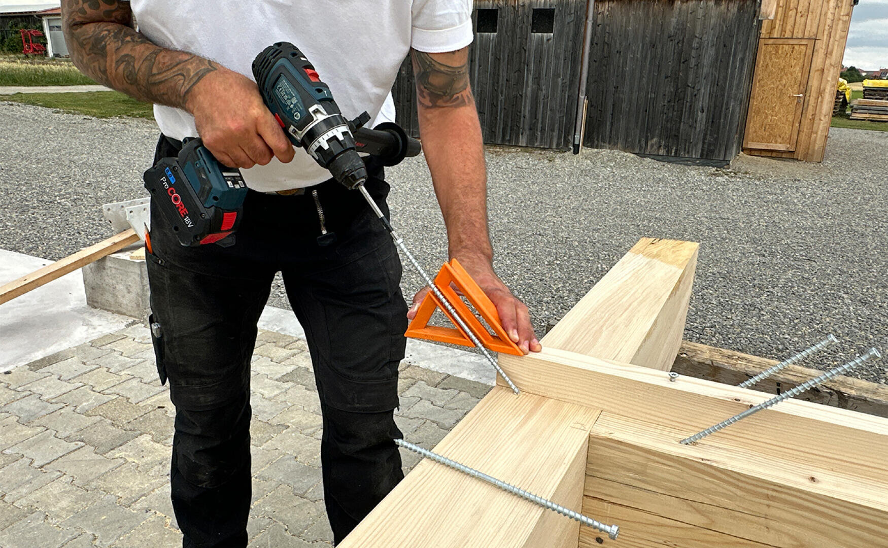 Eine Person setzt mit einer Bohrmaschine eine Schraube in einen Holzbalken ein und hält dabei ein orangefarbenes Zimmermannswinkel für präzise Messungen auf einer Baustelle im Freien.