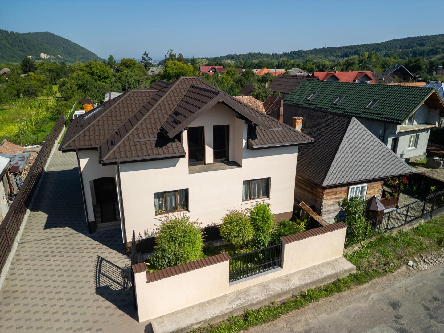 Ein modernes zweistöckiges Haus mit braunem Dach, cremefarbenen Wänden und kleinen grünen Sträuchern davor, neben einer gepflasterten Auffahrt und Nachbarhäusern, umgeben von Bäumen und Hügeln im Hintergrund.