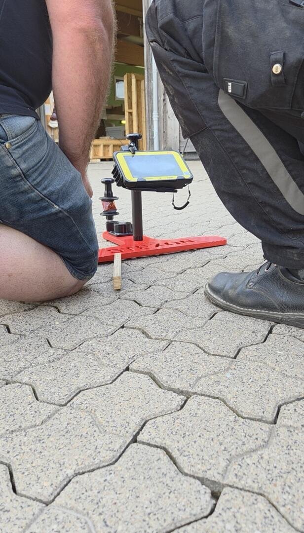 Zwei Personen knien auf ineinandergreifenden Pflastersteinen neben einem roten Gerät mit einer montierten Tafel und anderen Ausrüstungsgegenständen, möglicherweise für Vermessungsarbeiten. Im Hintergrund sind Paletten und eine Holzkonstruktion zu sehen.