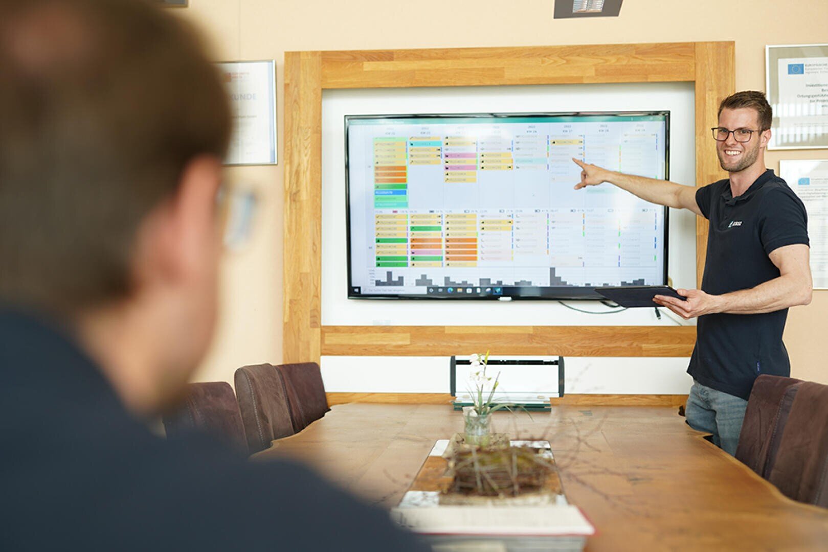 Ein Mann steht in einem Besprechungsraum und zeigt auf einen großen Bildschirm, auf dem bunte Datendiagramme angezeigt werden, während eine andere Person am Konferenztisch sitzt und aufmerksam zuhört.
