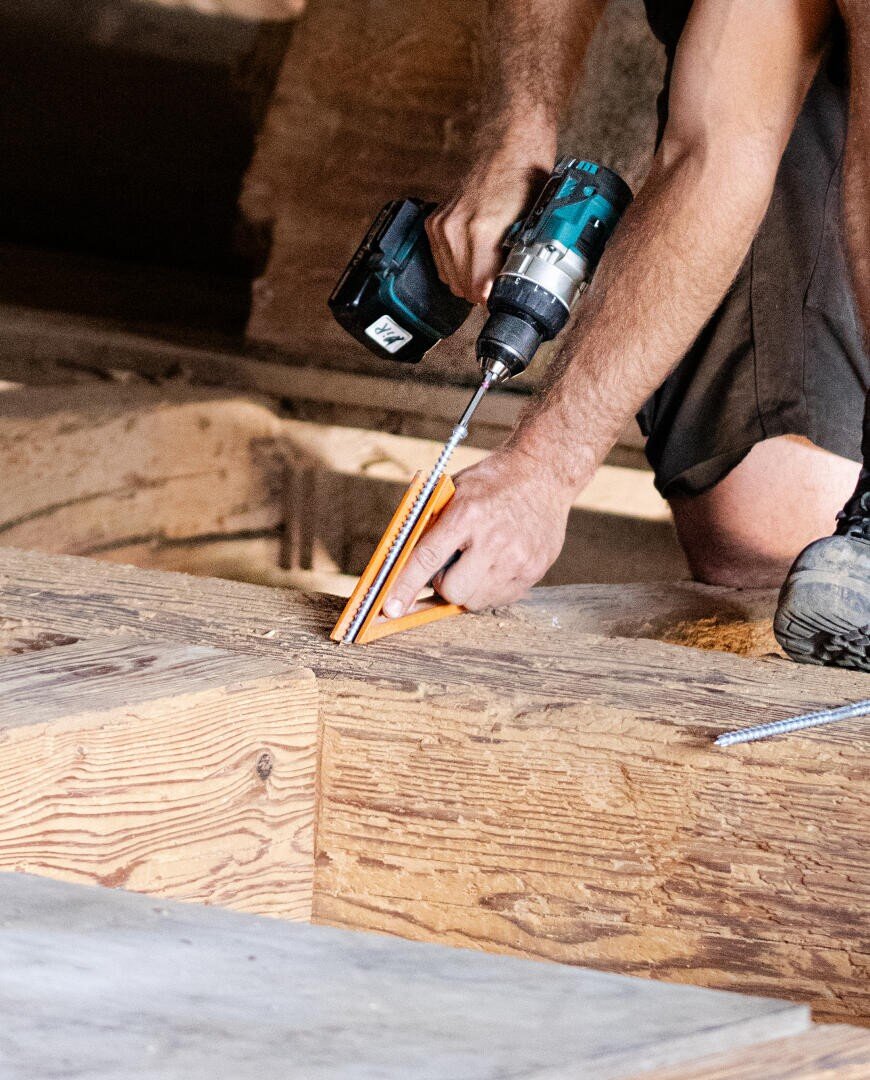 Eine Person verwendet einen Akku-Bohrschrauber und eine orangefarbene Führung, um auf einer Baustelle eine Schraube in eine Holzplanke zu bohren. Nur ihre Arme und Beine sind sichtbar.