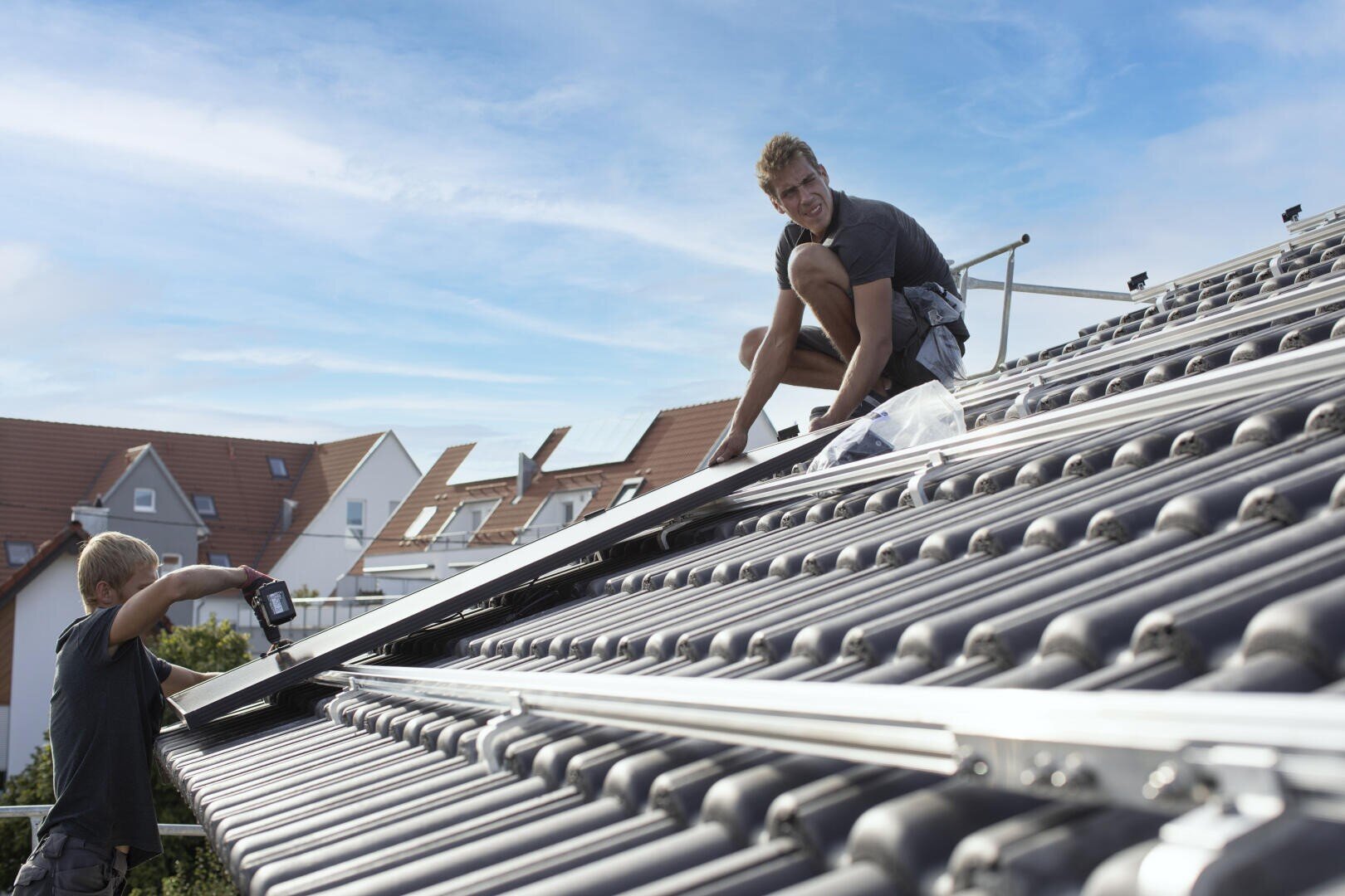 Zwei Arbeiter montieren bei klarem Himmel Metallschienen auf einem Ziegeldach, um die Installation von Solarmodulen vorzubereiten; im Hintergrund sind Häuser mit roten Dächern zu sehen.