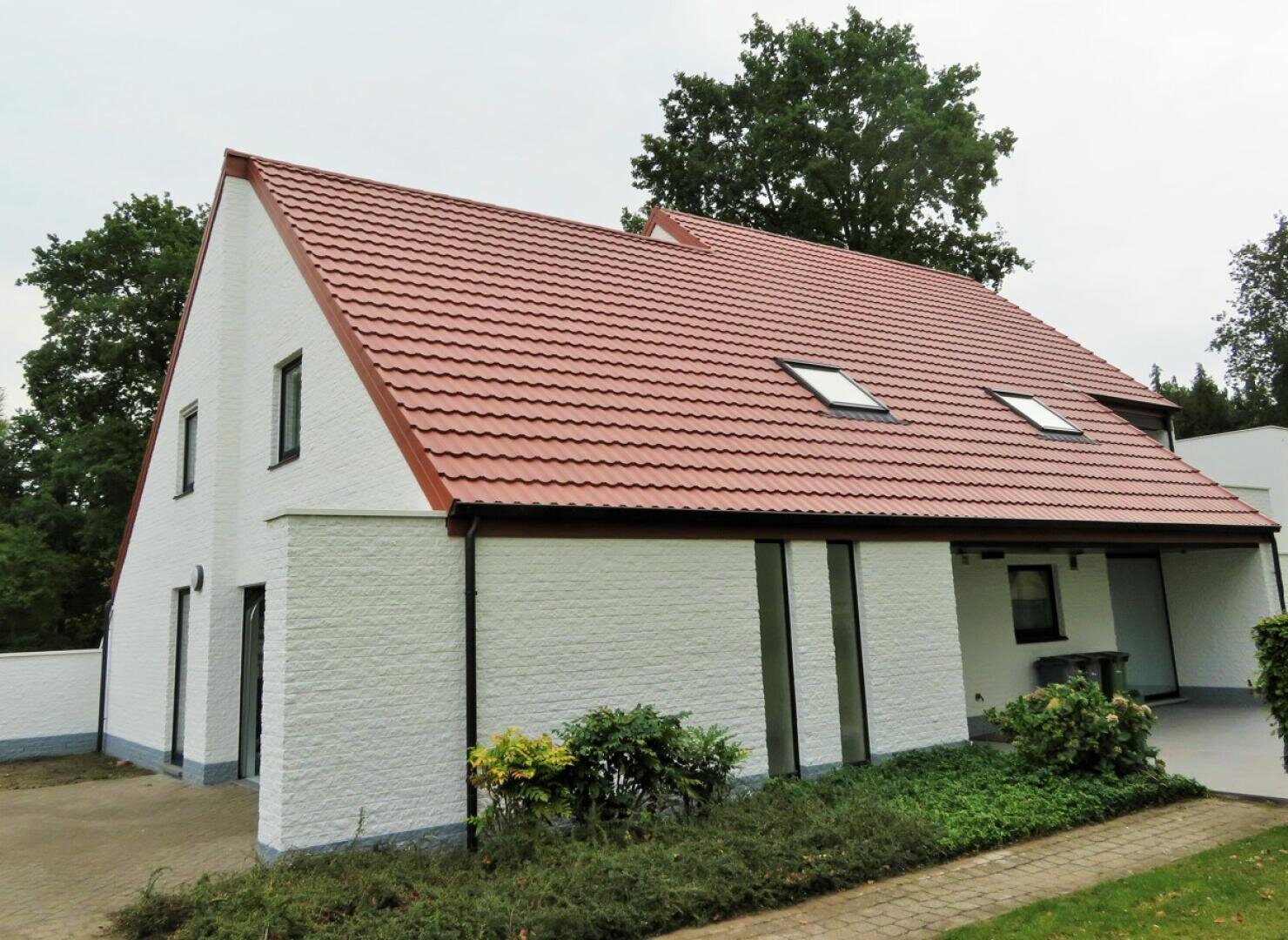 Ein modernes weißes Haus mit rotem Ziegeldach, Oberlichtern und mehreren Fenstern, umgeben von Grün und Bäumen. Ein gepflasterter Gehweg führt zum Eingang auf der rechten Seite.