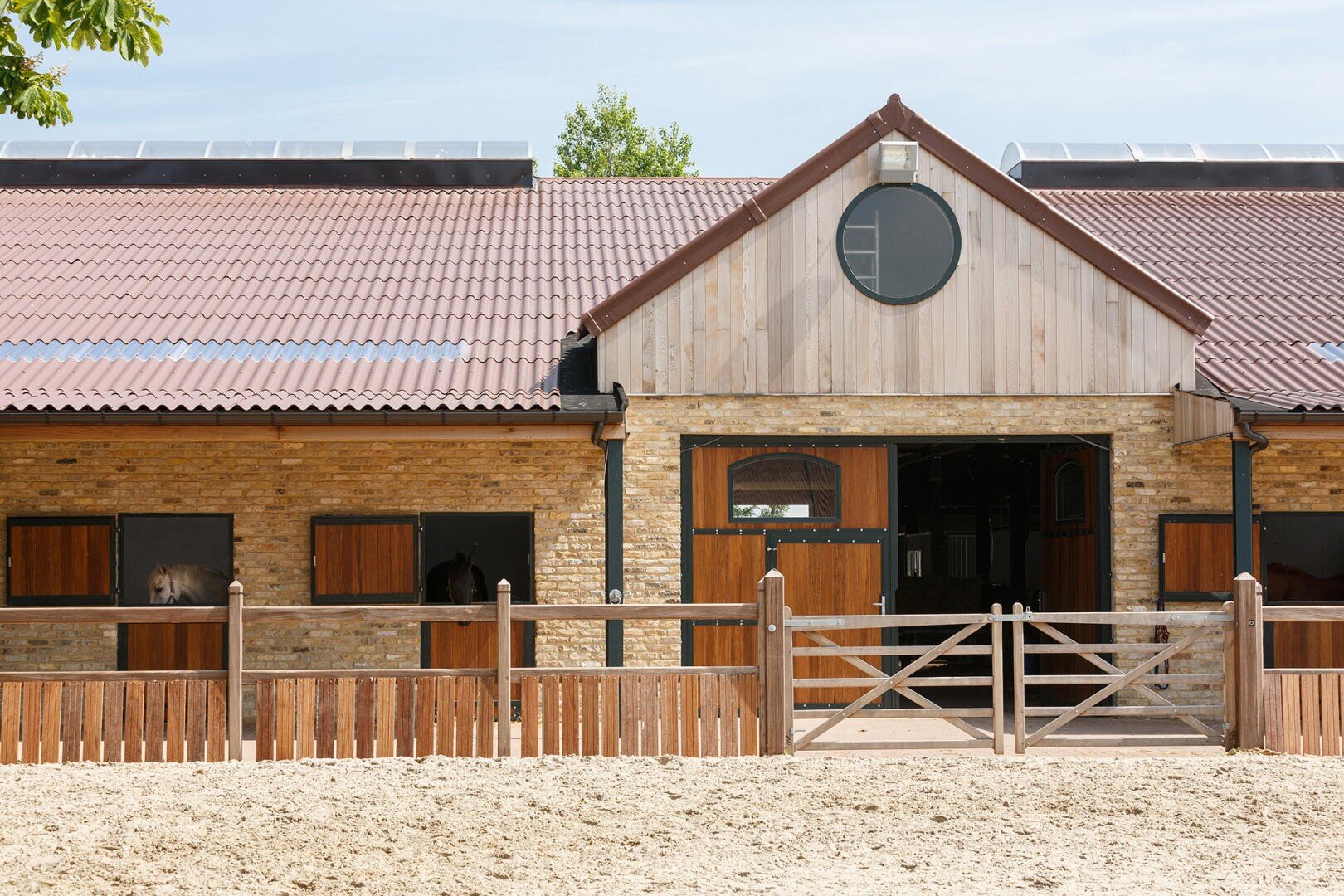 A modern brick and wood stable with several open wooden stall doors, some with horses inside, surrounded by a wooden fence and sandy ground under a clear sky.