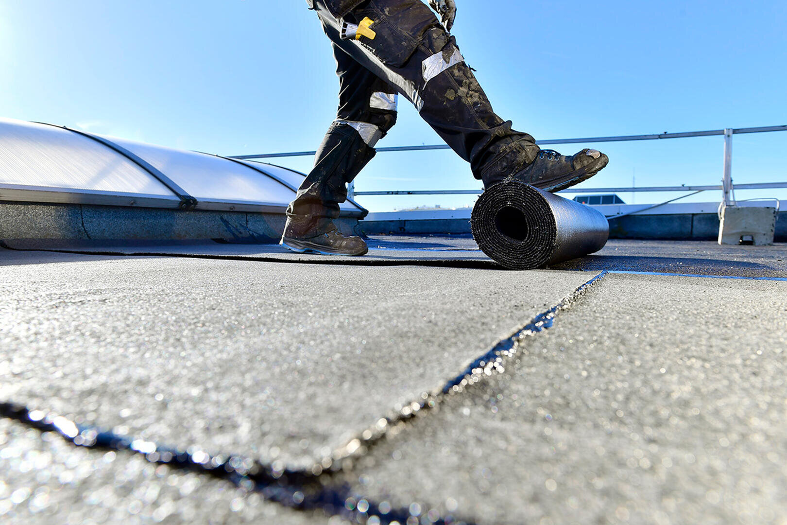 A worker in protective clothing rolls out a roofing membrane on a flat roof under a bright blue sky. In the background, tools are attached to his belt and safety rails can be seen.
