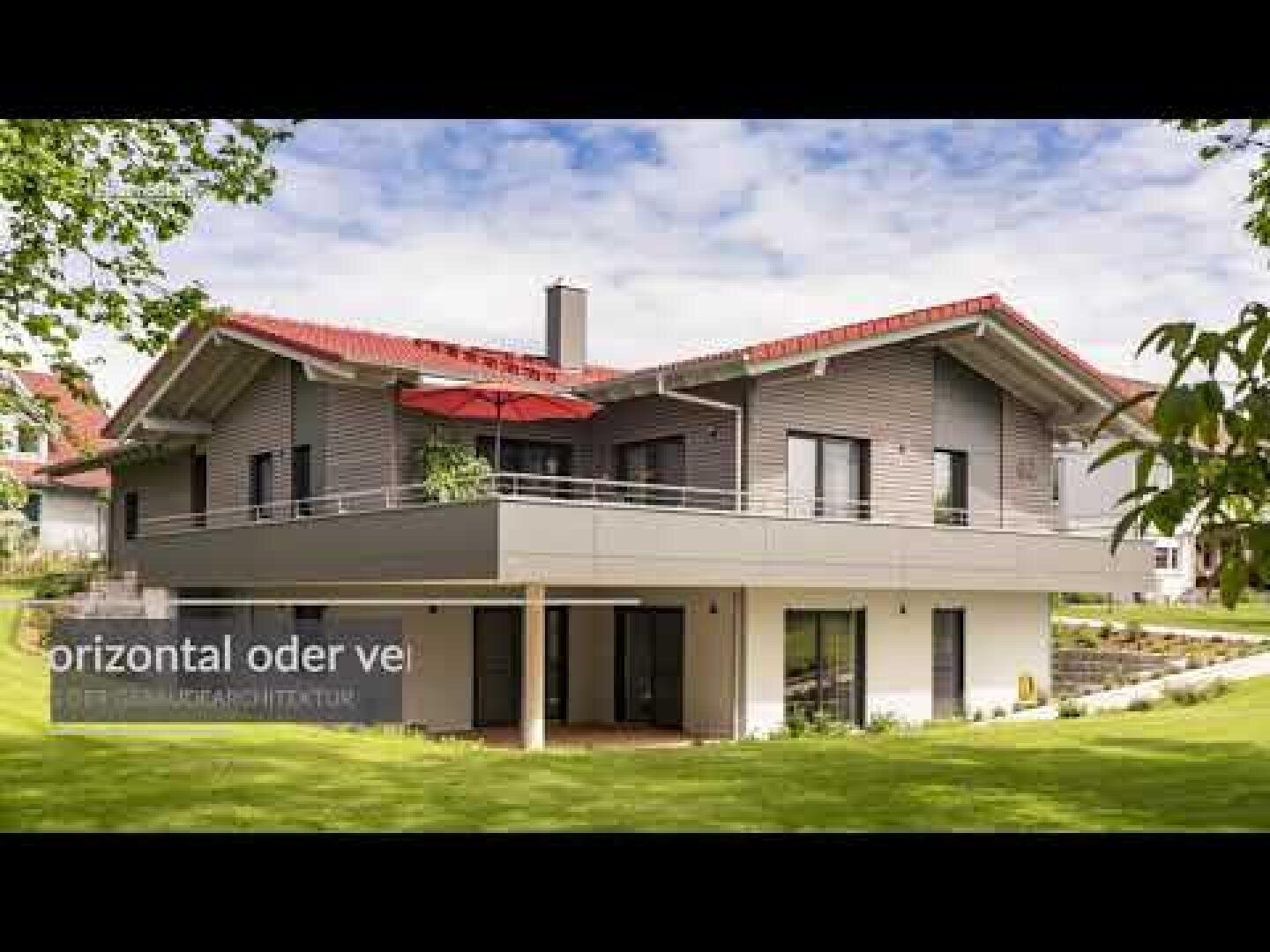 Modernes zweistöckiges Haus mit grauem Äußeren und rotem Ziegeldach, großen Fenstern, einem geräumigen Balkon mit rotem Sonnenschirm und einem gepflegten Rasen unter einem teilweise bewölkten Himmel.