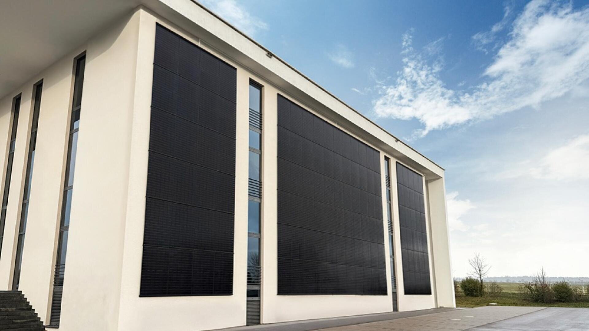 Ein modernes weißes Gebäude mit großen, vertikalen schwarzen Solarpaneelen, die den größten Teil der Außenwände bedecken, unter einem blauen Himmel mit vereinzelten Wolken. Ein gepflasterter Gehweg verläuft entlang des Gebäudes.