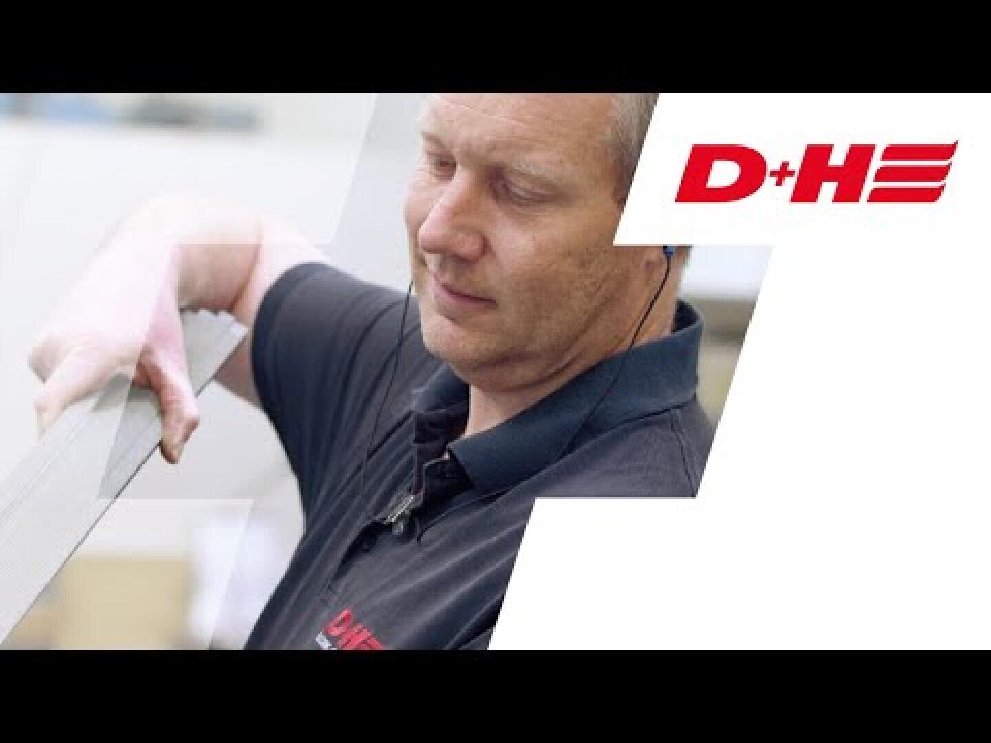 A man in a black polo shirt with a red D+H logo examines a long metal object. The background is predominantly white, with a large red D+H logo on the right-hand side.