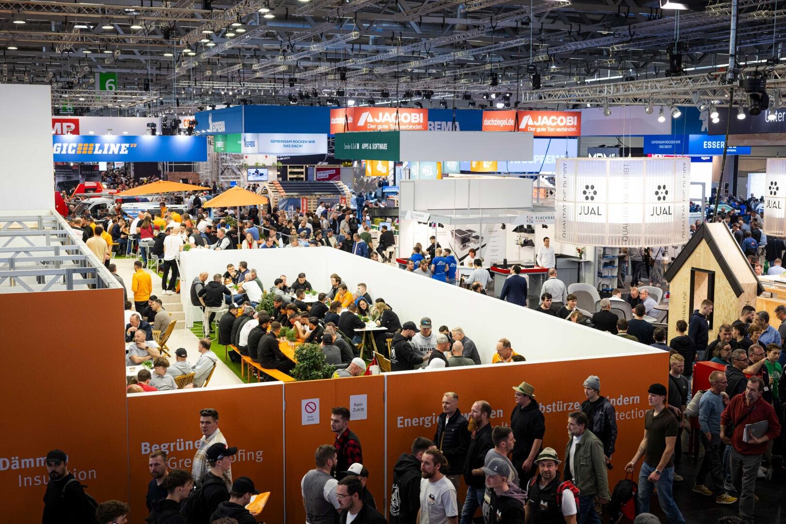 Eine belebte Messehalle mit vielen Menschen, die um die Stände herumgehen und an Tischen in einem weißwandigen Sitzbereich sitzen. Bunte Banner und Firmenschilder hängen über der überfüllten Messehalle.