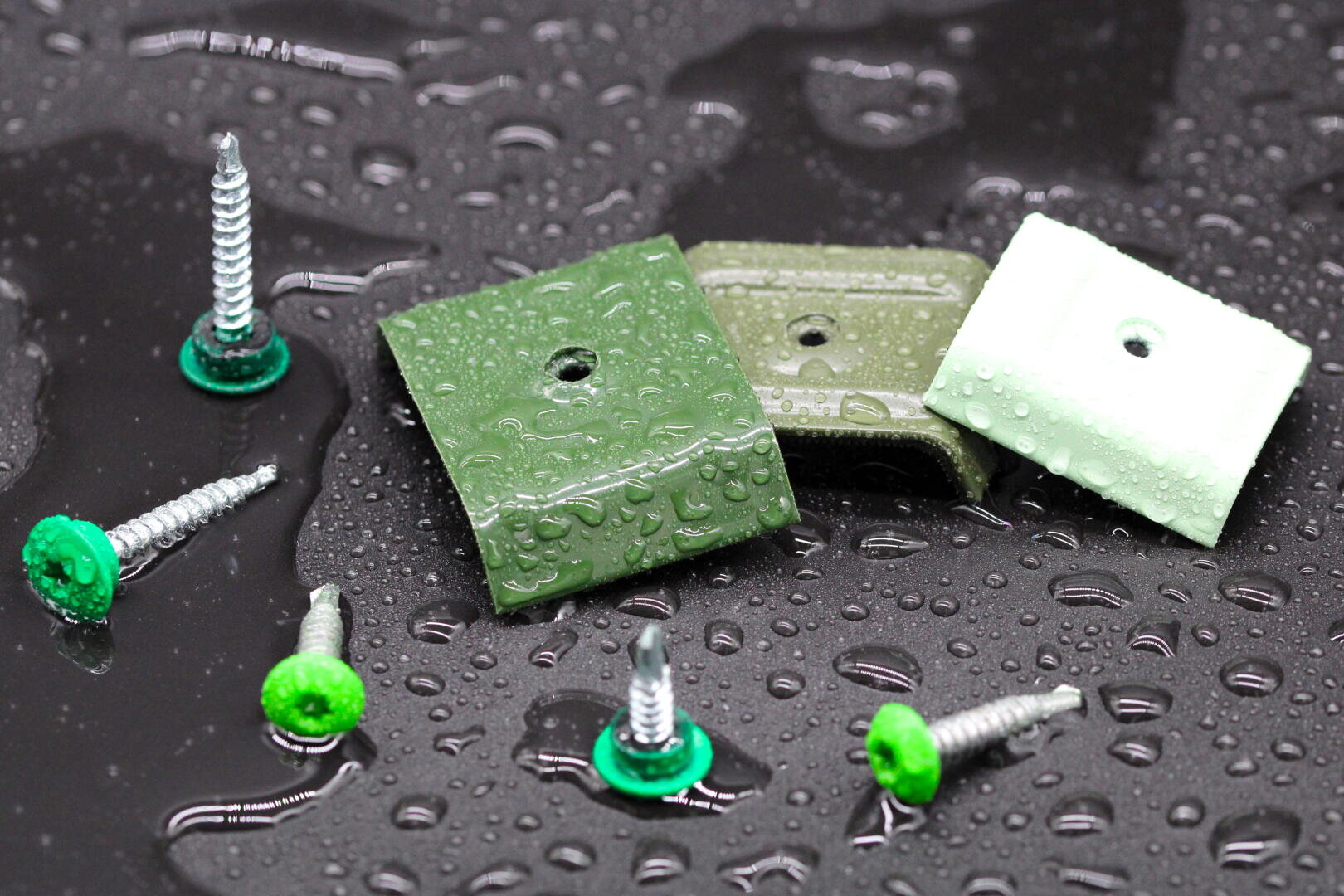 Drei grüne Metallbügel und sechs Schrauben mit grünen Unterlegscheiben liegen auf einer nassen, dunklen Oberfläche, die mit Wassertropfen bedeckt ist.