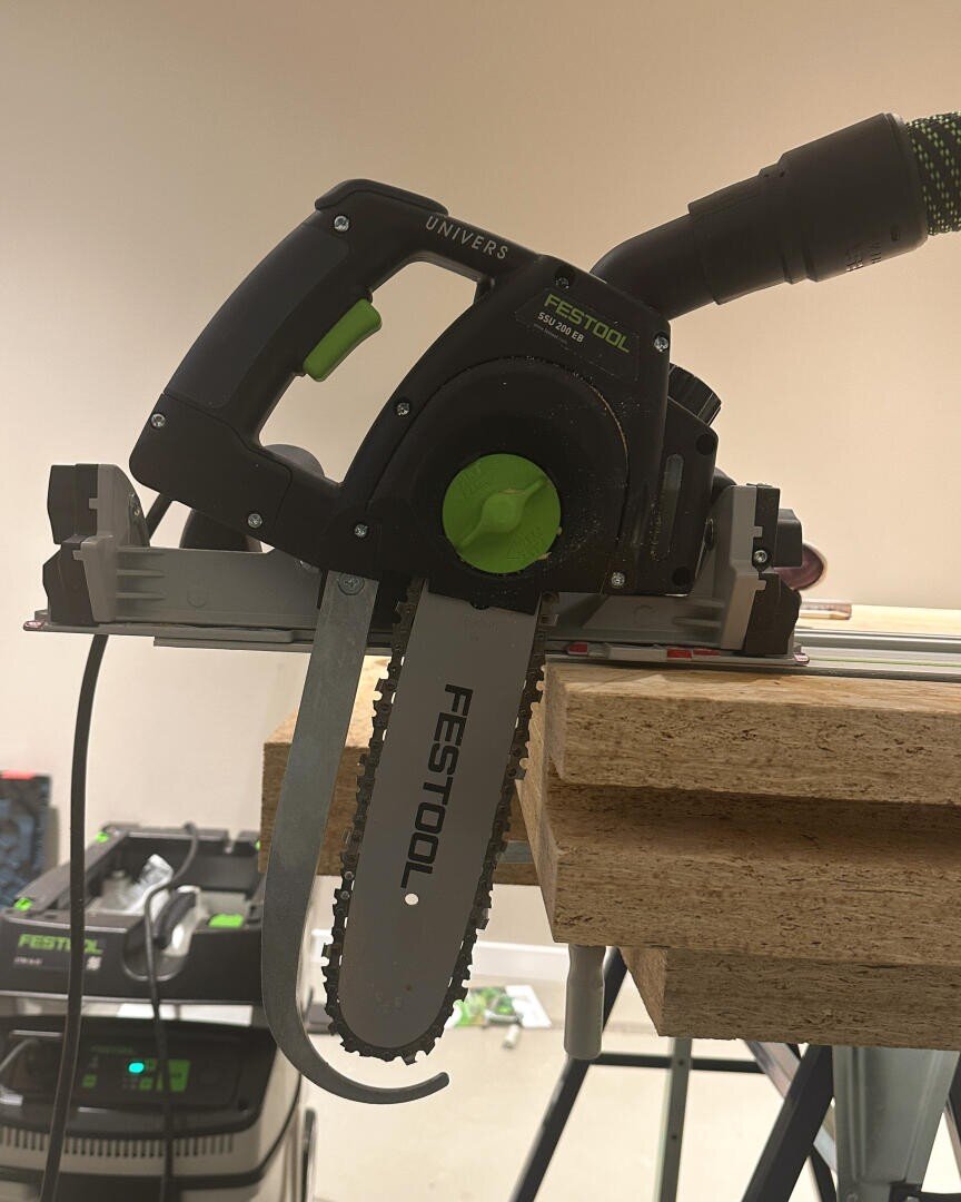 Eine Festool Kettenfräse steht auf einem Stapel dicker Holzbretter, das Kettenblatt ist zum Schneiden senkrecht aufgestellt, und ein Absaugschlauch ist angebracht. Im Hintergrund sind Werkstattausrüstung und Werkzeuge zu sehen.