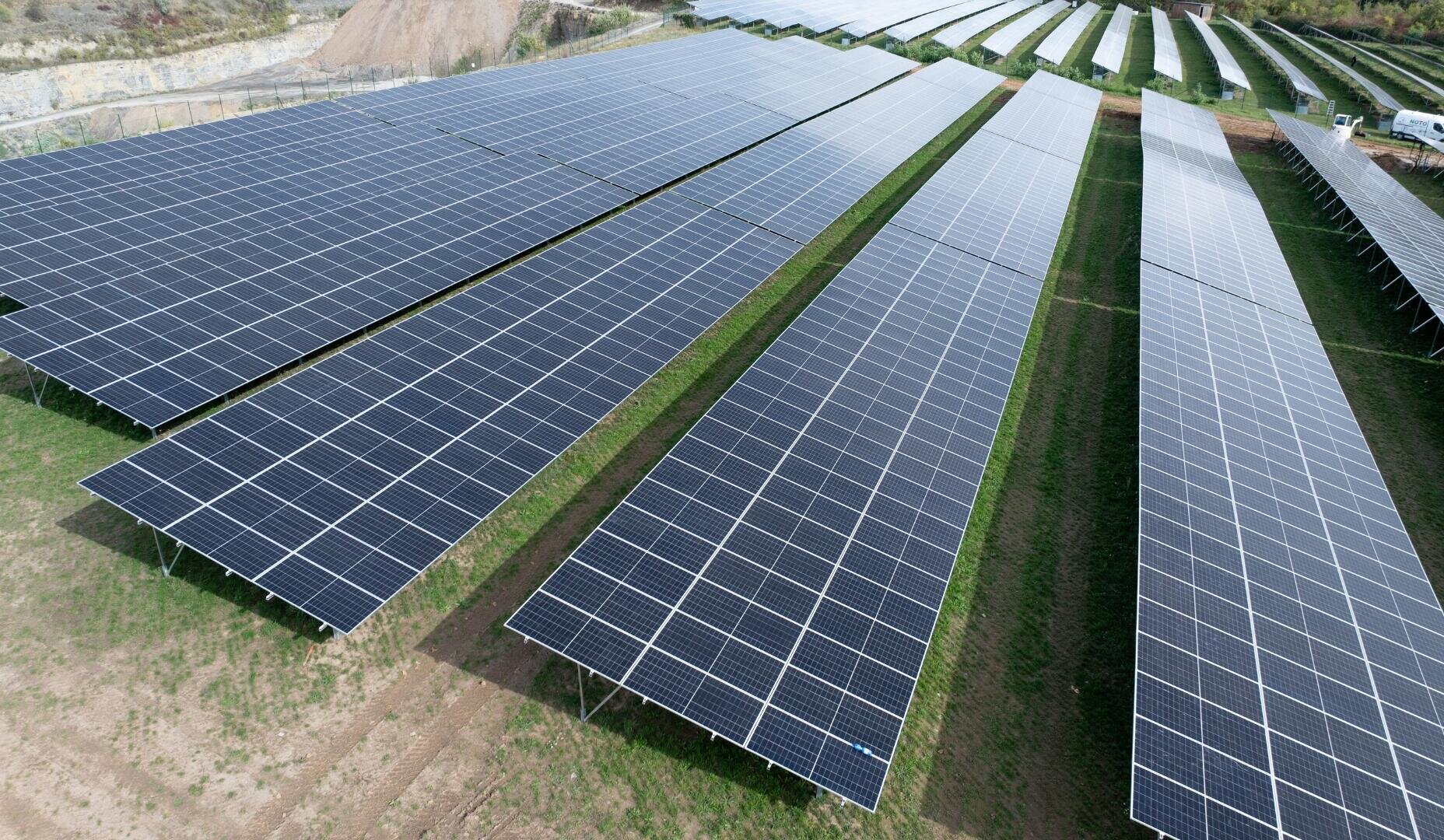 Reihen von großen Solarmodulen, die auf einer Wiese installiert sind und einen Solarpark bilden. Die Paneele sind so ausgerichtet, dass sie das Sonnenlicht einfangen, während im Hintergrund Bäume und Industrieanlagen zu sehen sind.