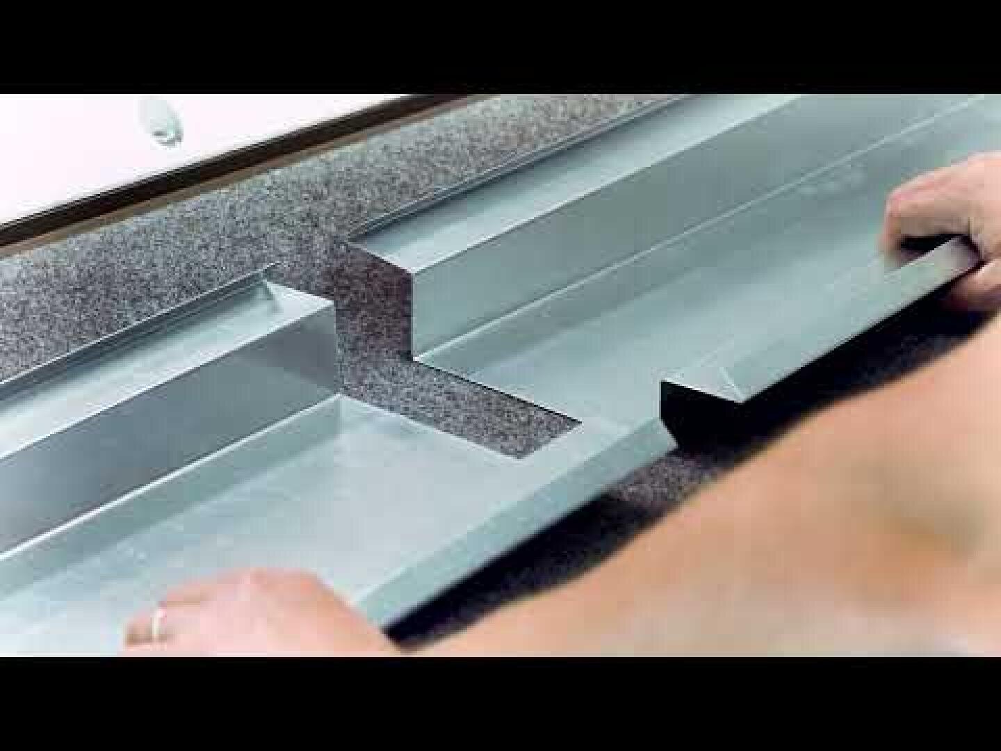 A person positions two pieces of metal flashing with interlocking notches on a gray surface, preparing to join them together.