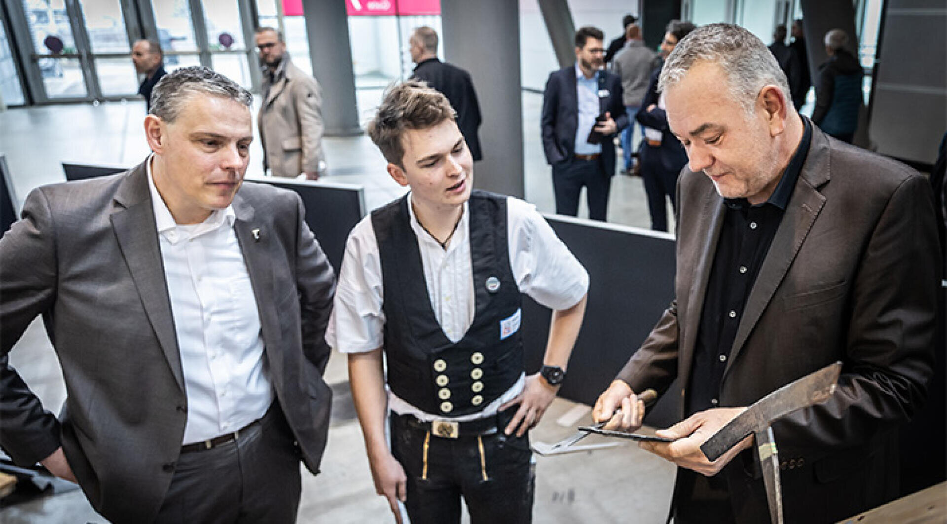 Drei Männer stehen in einem Innenraum bei einer Veranstaltung zusammen. Zwei Männer in Anzügen beobachten, während ein dritter Mann, der eine Weste und Freizeitkleidung trägt, mit einem Metallgegenstand in der Hand etwas demonstriert oder erklärt. Im Hintergrund sind weitere Personen zu sehen.