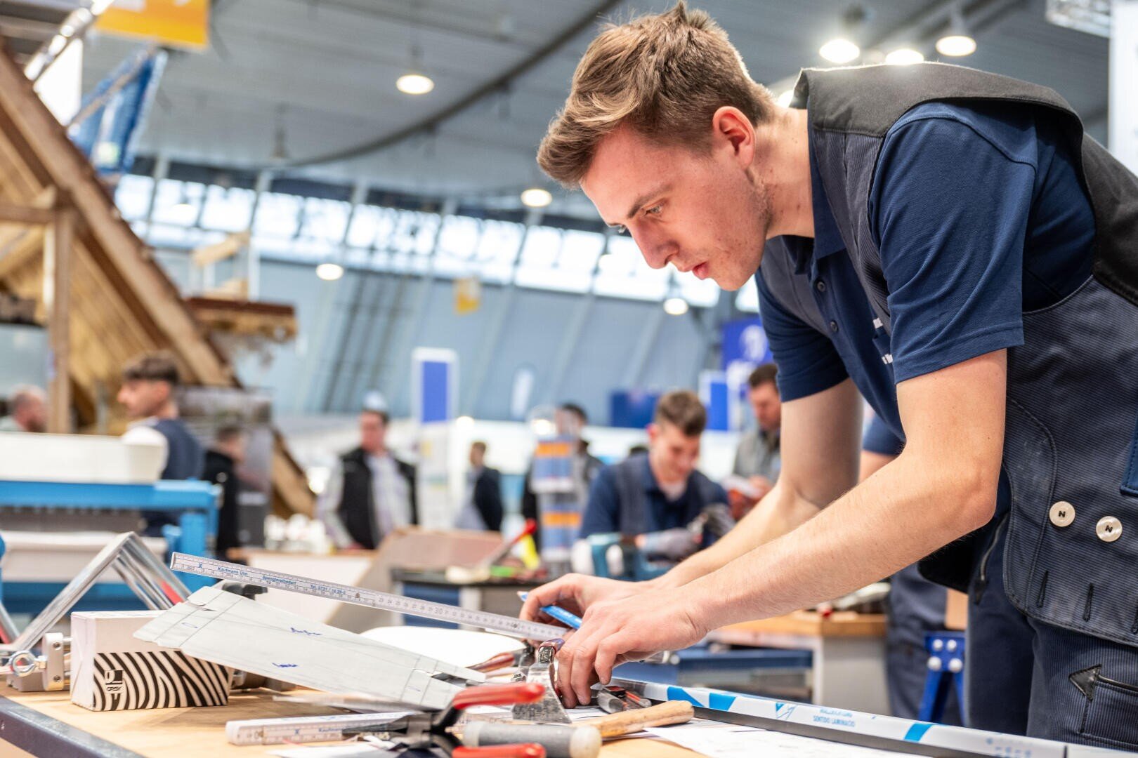 Ein junger Mann in einer blauen Weste misst und markiert ein Stück Material auf einem Tisch mit verschiedenen Werkzeugen, konzentriert auf seine Arbeit in einer hell erleuchteten, geschäftigen Werkstatt oder Industrieumgebung.