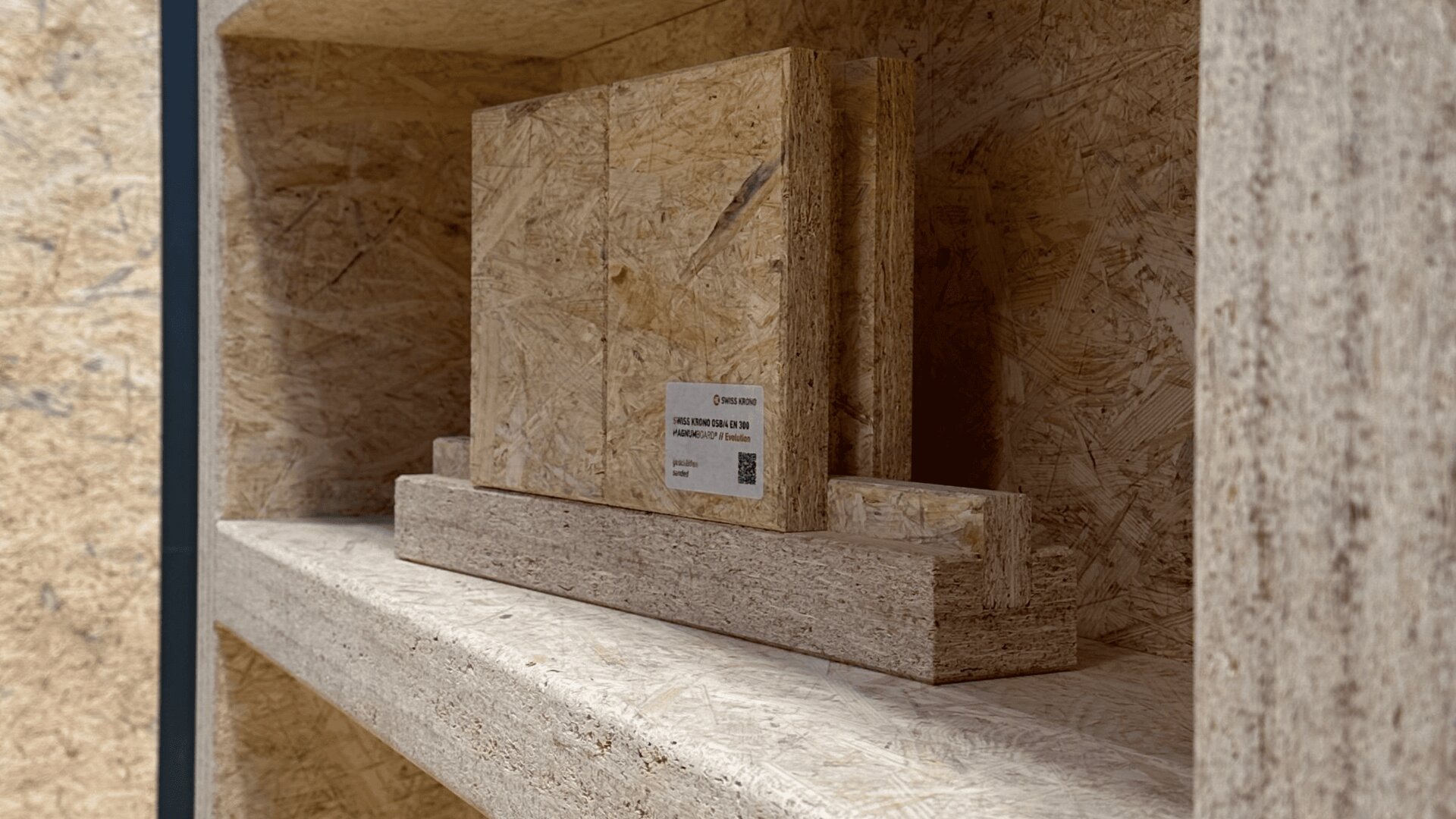 Nahaufnahme eines Holzregals mit gestapelten OSB- und Spanplattenproben. An einer der Proben ist ein kleines Etikett angebracht. Regal und Platten wirken rau und strukturiert.