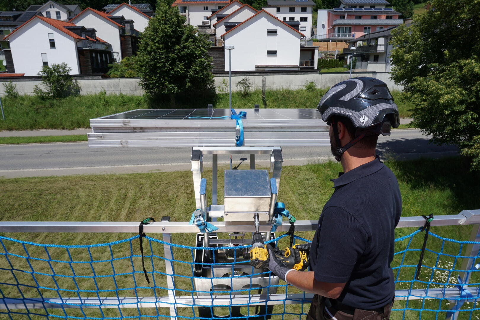 Eine Person mit Helm bedient ein Gerät neben einem Sicherheitsnetz auf einer erhöhten Plattform, im Hintergrund sind Metallträger und eine Straße zu sehen, dahinter Häuser auf einem grasbewachsenen Hügel.