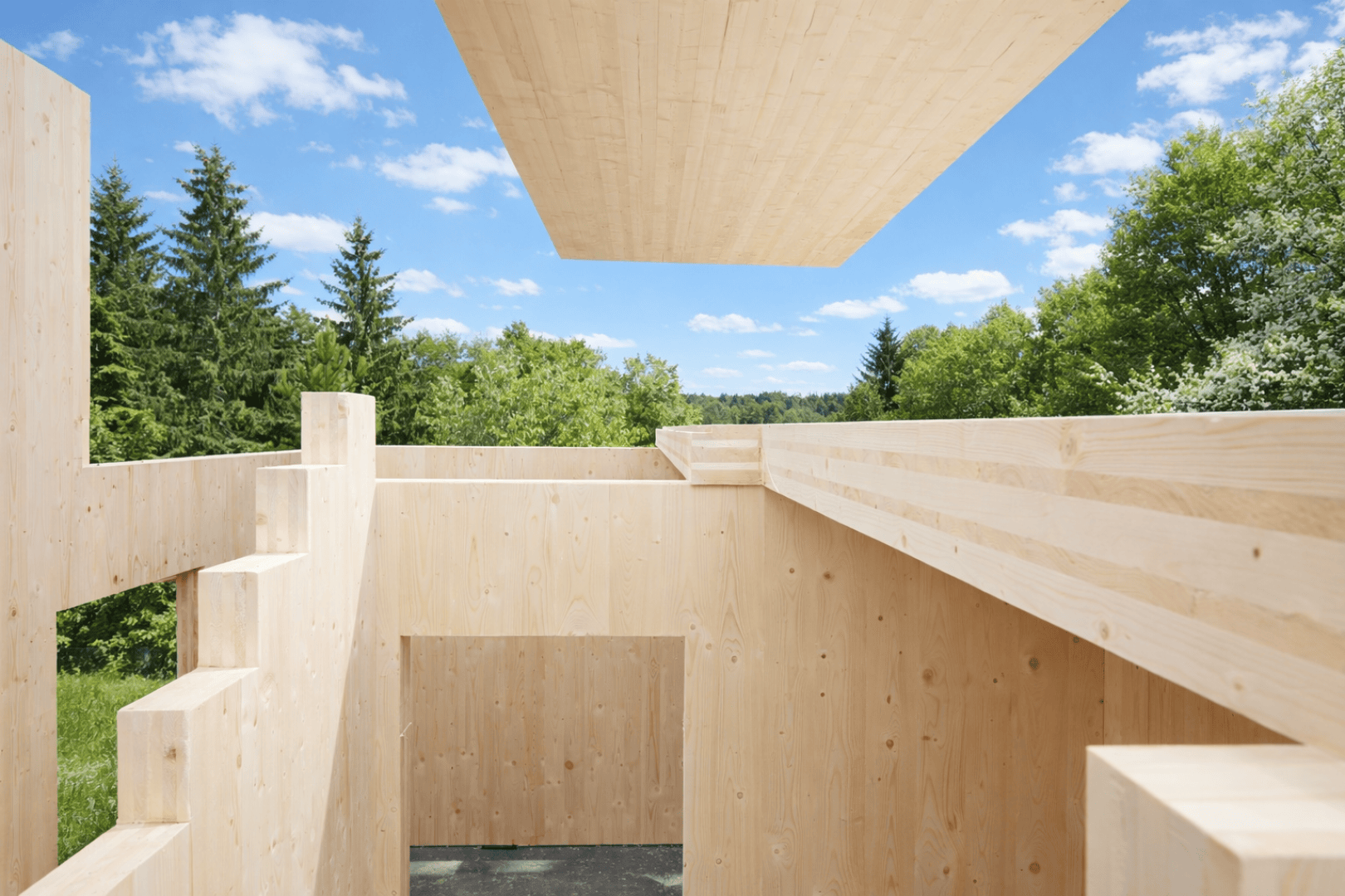 Ein modernes, im Bau befindliches Holzgebäude mit freiliegenden Wänden und Balken im Freien, umgeben von grünen Bäumen und einem blauen Himmel mit vereinzelten Wolken. Eine große Holzplanke wird über dem Bauwerk angebracht.