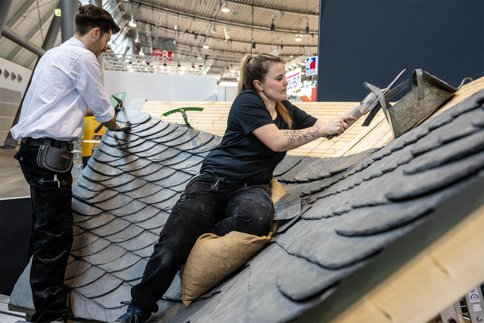 Two people are working on a pitched roof, fitting and fixing dark slate tiles. One is using a hammer and chisel while kneeling on a cushion and the other is fitting the tiles. They are in a large, well-lit building.