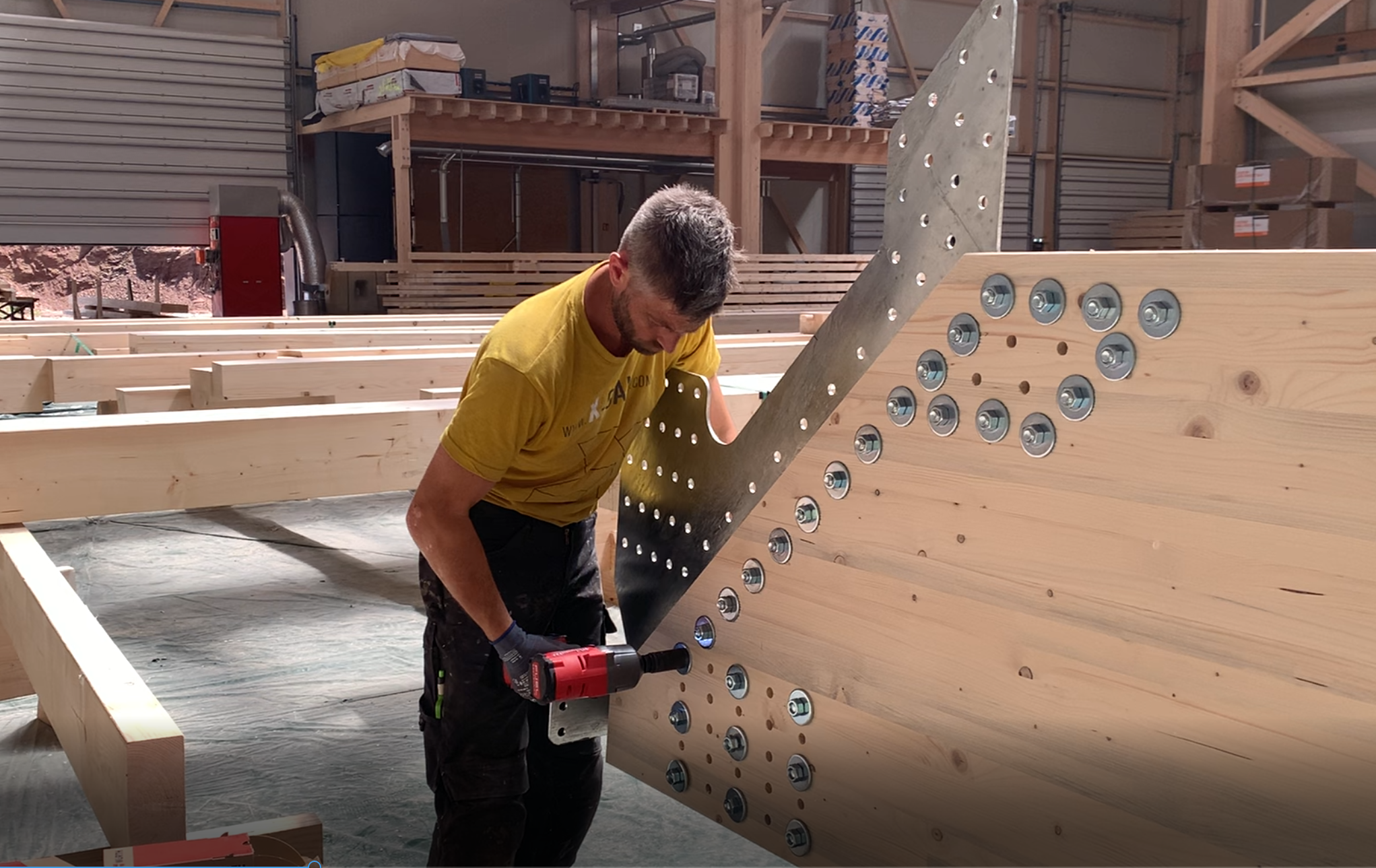 Ein Mann in einem gelben Hemd benutzt eine Bohrmaschine, um Metallklammern an gro&szlig;en Holzbalken in einer Werkstatt oder auf einer Baustelle zu befestigen.