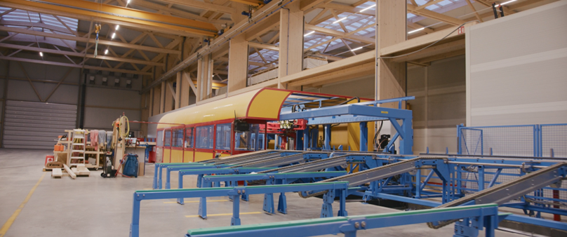 Ein Fabrikinterieur mit einem gelben und roten Stra&szlig;enbahn- oder Eisenbahnwagen, der zusammengebaut wird, umgeben von blauen F&ouml;rderanlagen, Werkzeugen und Lagerr&auml;umen in einem ger&auml;umigen, gut beleuchteten Geb&auml;ude.
