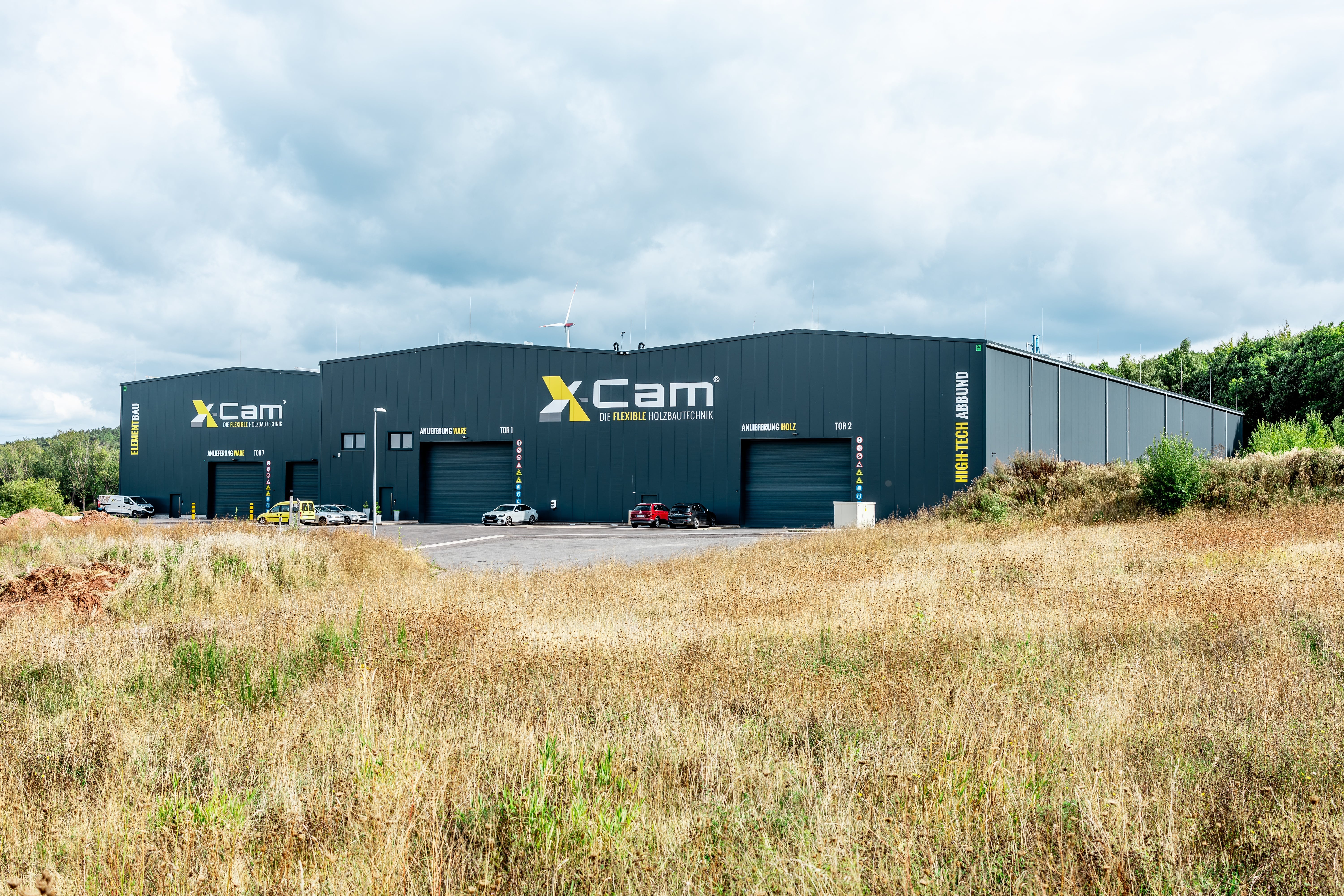Ein gro&szlig;es Industriegeb&auml;ude mit dem Xcam-Schriftzug an der Au&szlig;enseite steht inmitten von grasbewachsenen Feldern unter einem bew&ouml;lkten Himmel. Vor dem Geb&auml;ude sind mehrere Autos geparkt, und im Hintergrund begrenzen B&auml;ume die Szene.