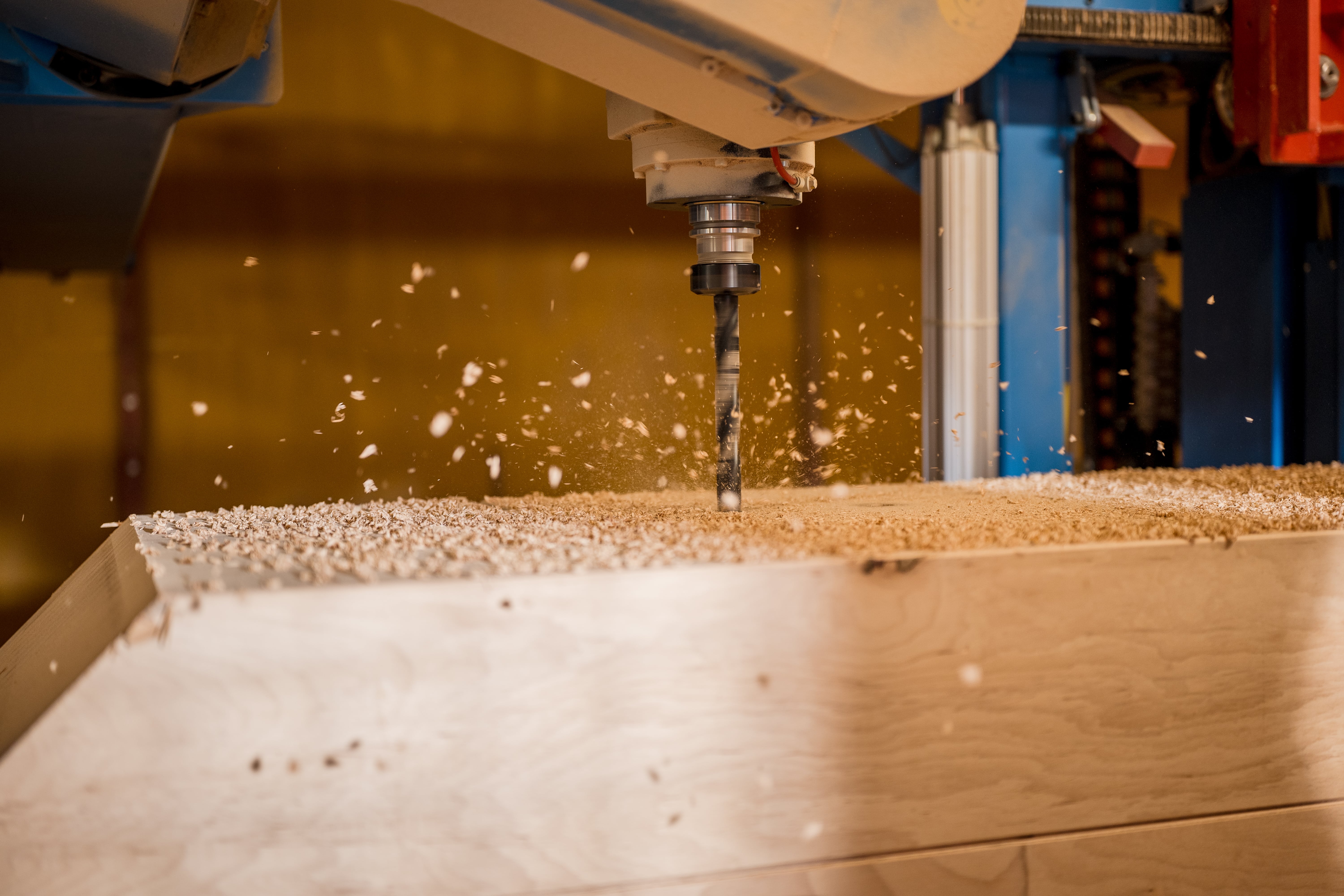Nahaufnahme einer CNC-Maschine, die in ein Holzbrett bohrt, wobei Holzsp&auml;ne und Staub in der Luft fliegen. Der Hintergrund ist leicht unscharf, um die Bewegung und die Pr&auml;zision des Bohrvorgangs hervorzuheben.