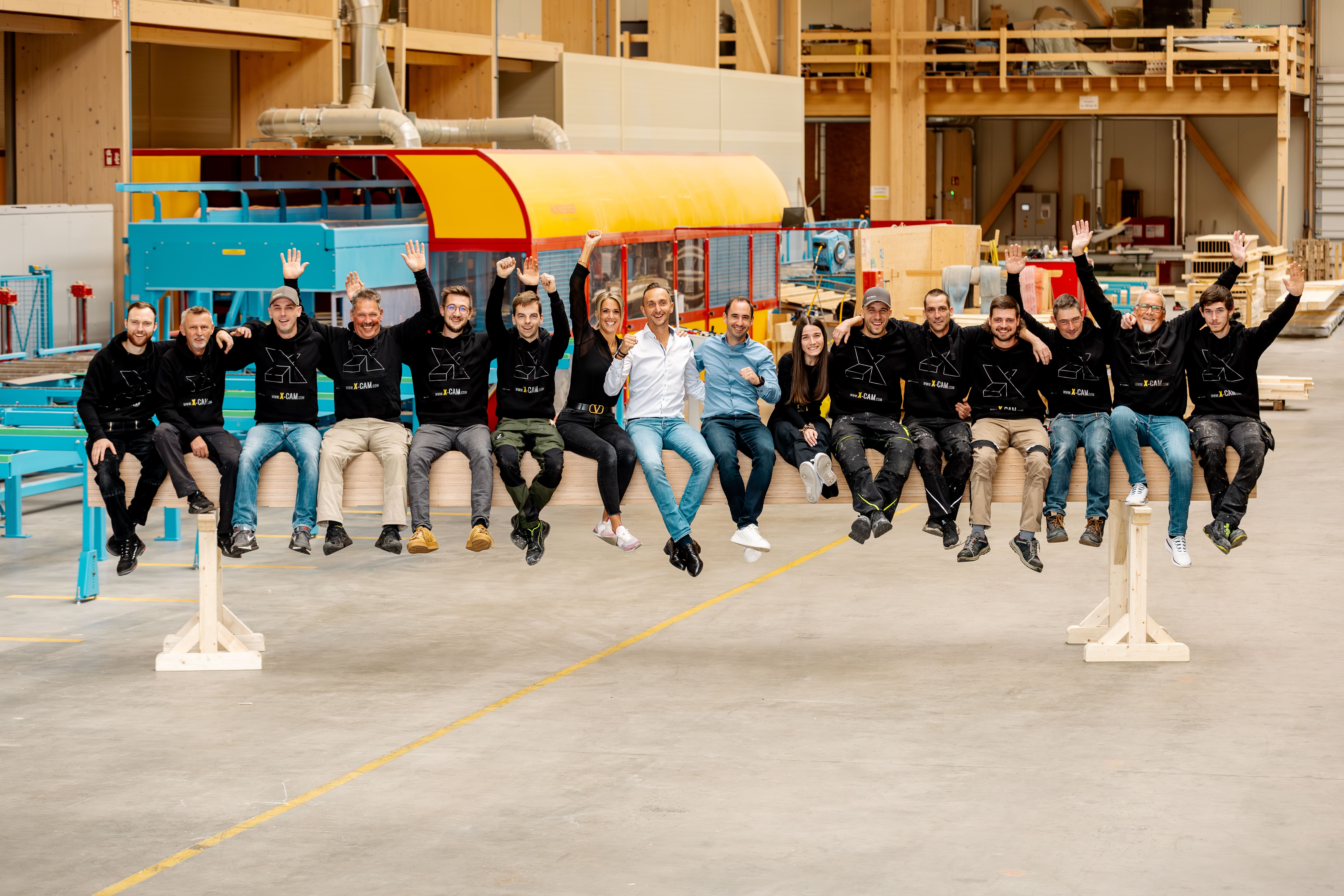 Eine Gruppe von 16 Personen sitzt auf einem Holzbalken in einer gro&szlig;en Werkstatt, l&auml;chelt und winkt in die Kamera. Die meisten tragen passende schwarze Kapuzenpullover; zwei Personen in der Mitte tragen wei&szlig;e Hemden. Hinter ihnen sind Baumaschinen zu sehen.