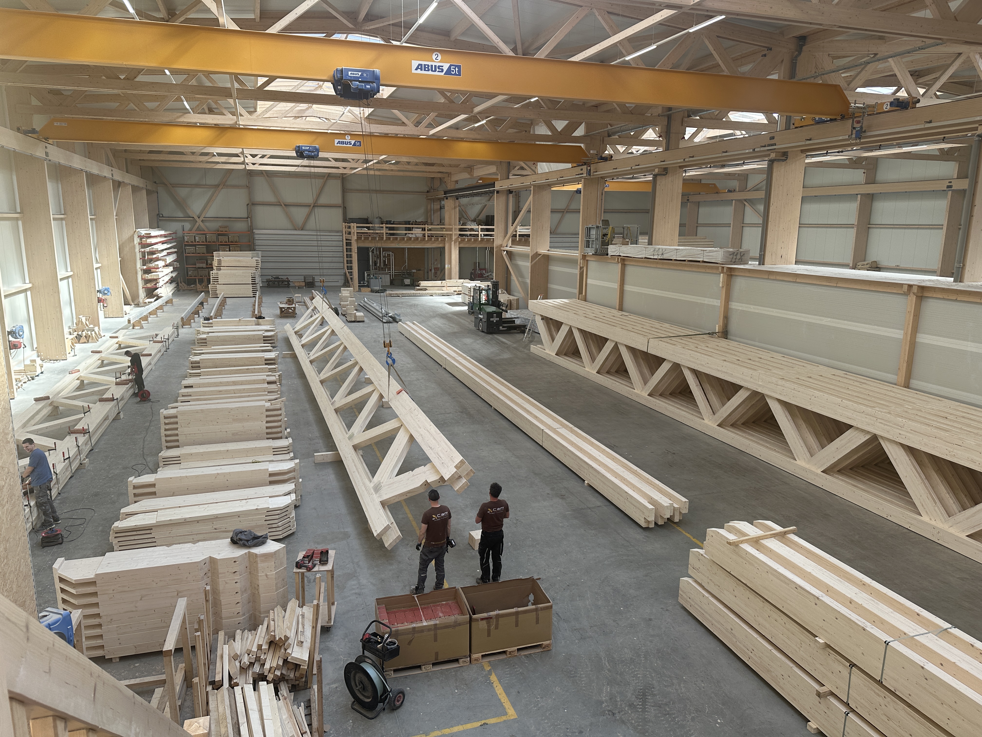 Eine geräumige Holzwerkstatt mit großen Holzbalken, gestapelten Brettern, Baumaschinen und zwei Arbeitern in kastanienbraunen Hemden, die neben einer langen Holzkonstruktion in der Mitte stehen.