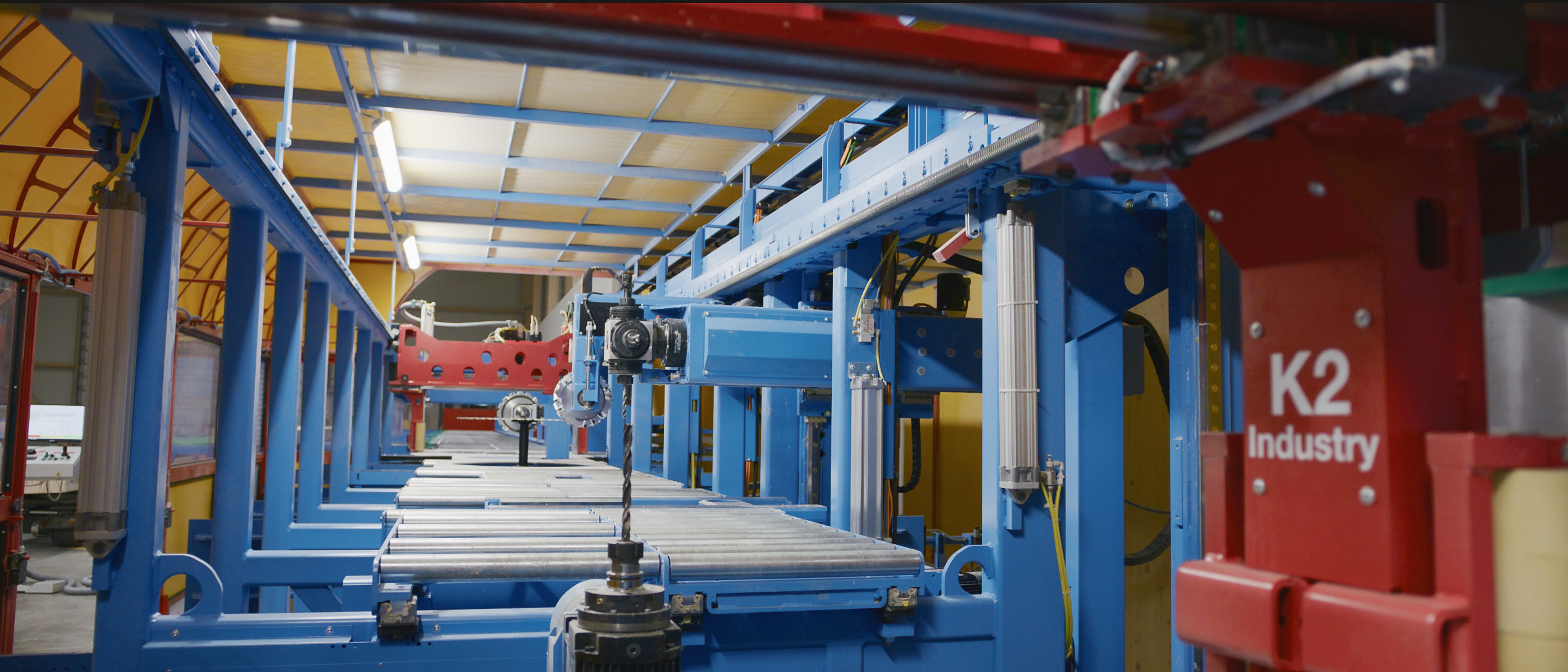 Nahaufnahme eines industriellen F&ouml;rdersystems mit blauen Metallrahmen, Rollen und Robotermaschinen, mit einer roten Tafel mit der Aufschrift K2 Industry auf der rechten Seite.