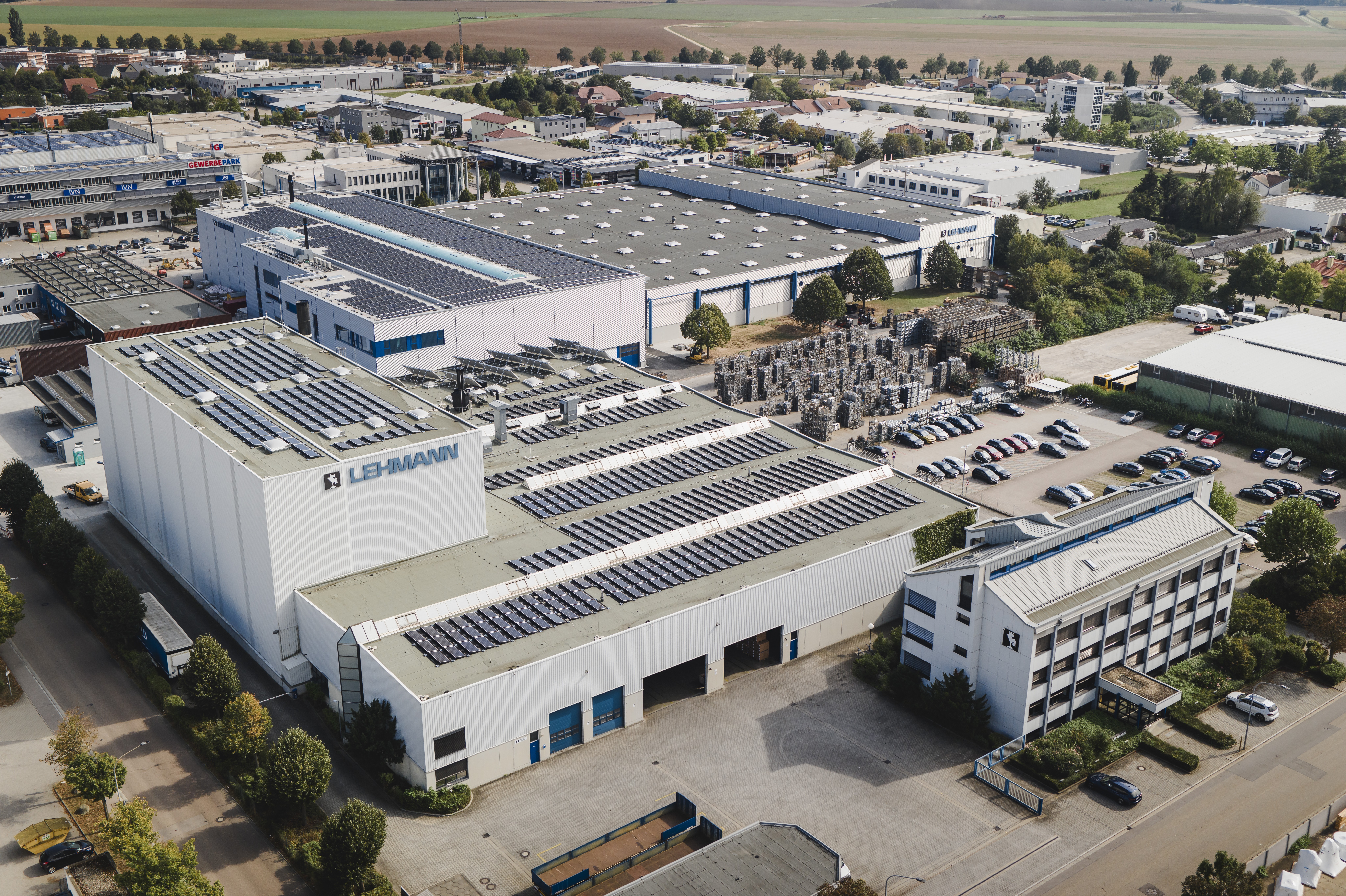 Luftaufnahme eines Industriekomplexes mit großen weißen Gebäuden, von denen viele Solarzellen auf ihren Dächern haben. Ein Parkplatz mit Autos, Bäume und umliegende Felder sind zu sehen. Auf dem Hauptgebäude steht der Name LEHMANN.