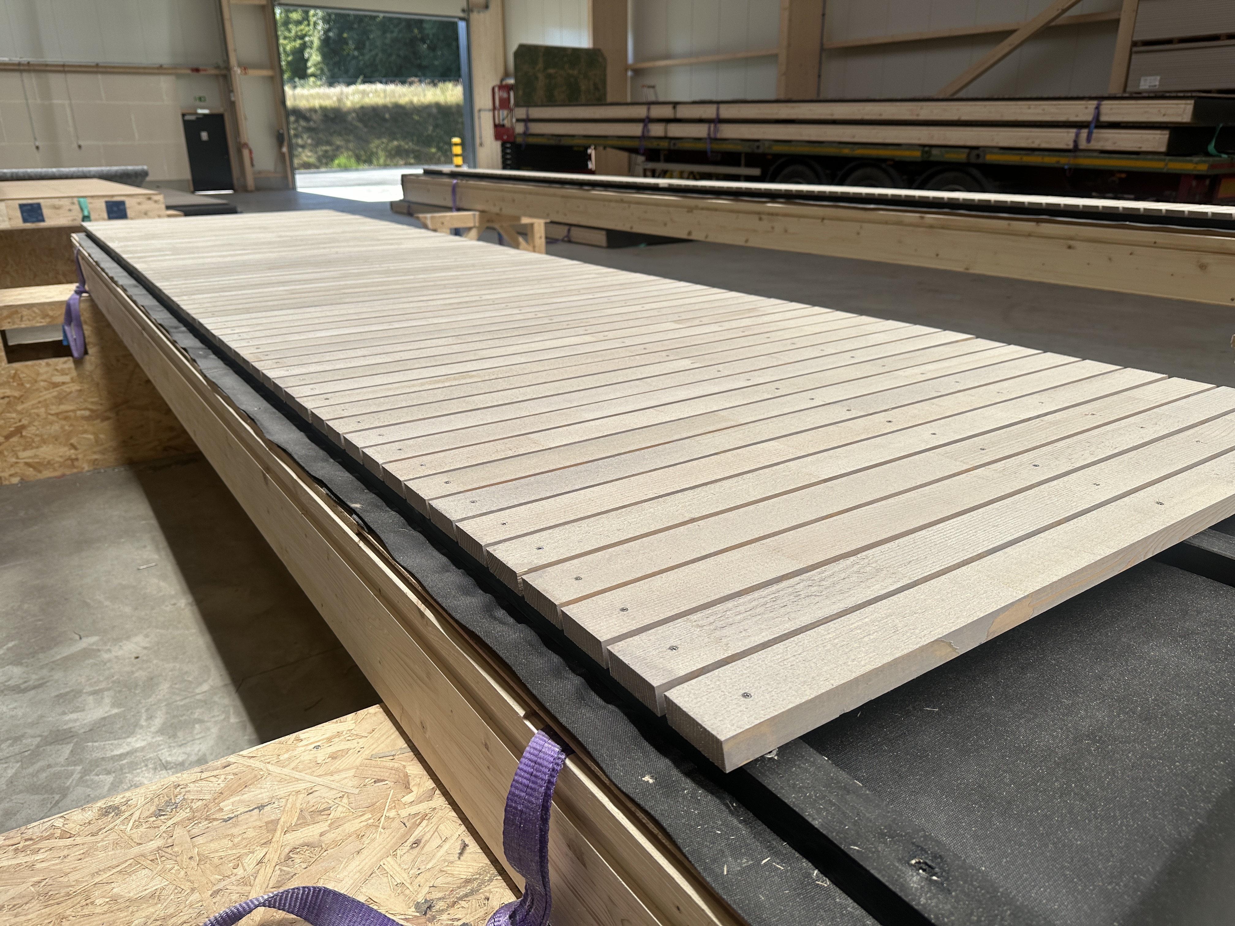 Nahaufnahme einer gro&szlig;en, rechteckigen Holzplatte mit gleichm&auml;&szlig;ig verteilten Lamellen, die auf einer Werkbank in einem gut beleuchteten Industrielager steht. Das Sonnenlicht str&ouml;mt durch offene T&uuml;ren im Hintergrund.