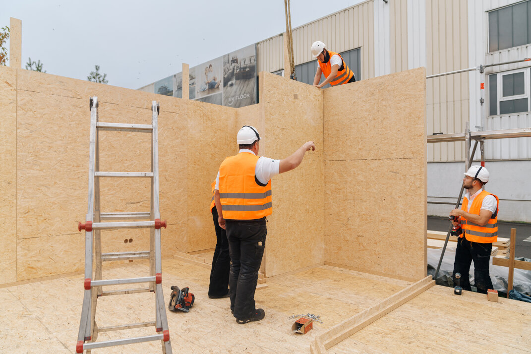 Four construction workers in orange safety vests and white helmets assemble wooden walls at a building site. A ladder and tools are visible on the plywood floor, and a large building is in the background.