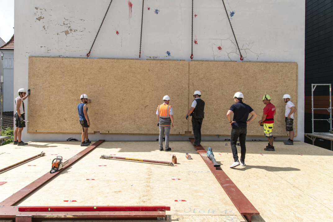 Seven construction workers in safety gear stand on a wooden platform, guiding a large prefabricated wall panel into place with the help of cranes. Tools and equipment are scattered on the platform.