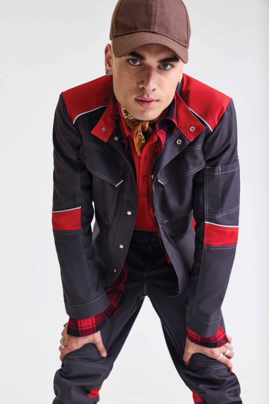 A person wearing a brown cap, a red and black jacket with red accents, a red shirt and a patterned scarf leans forward with his hands on his knees against a plain light background.