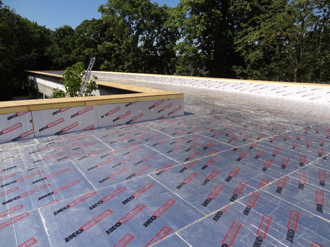 Flat roof under construction with sheets of insulation material labeled IKO Enertherm covering the surface; trees and a ladder are visible in the background.