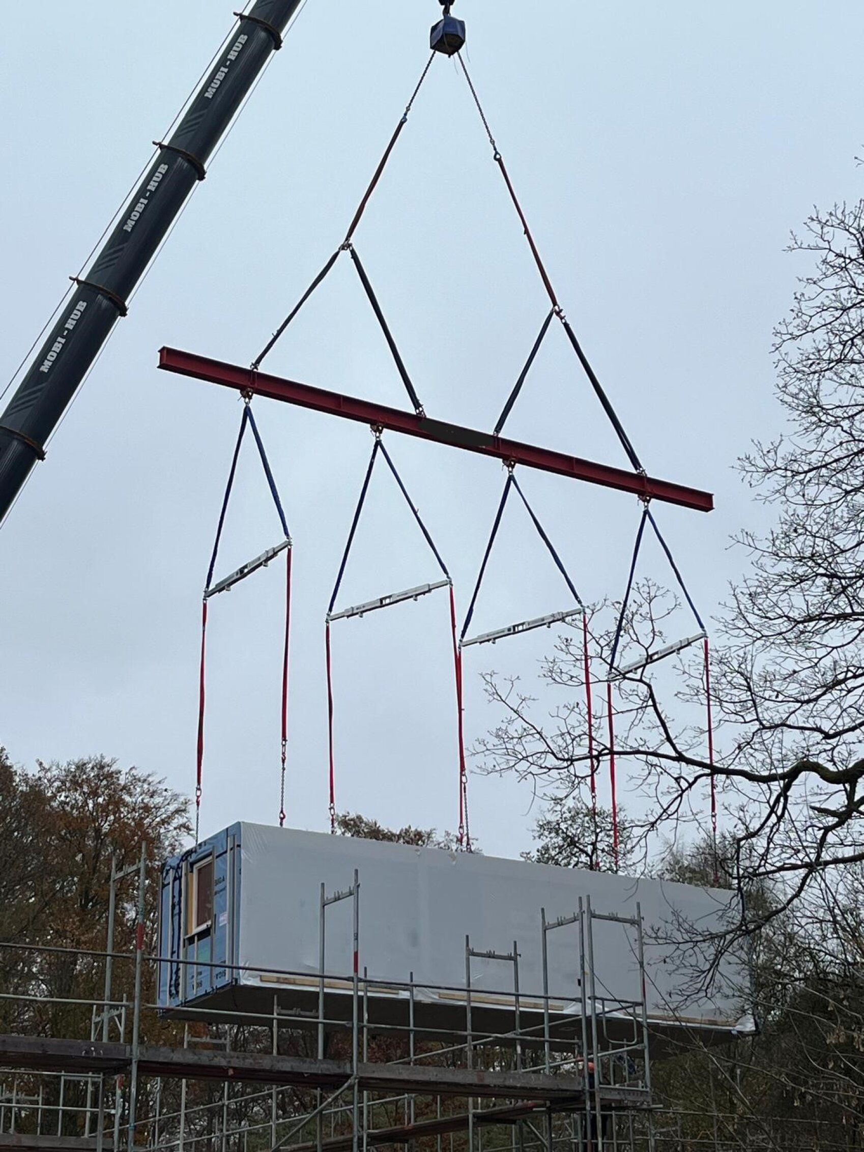A crane lifts a large rectangular prefabricated building module with red straps over scaffolding and trees to place it on a construction site under a cloudy sky.