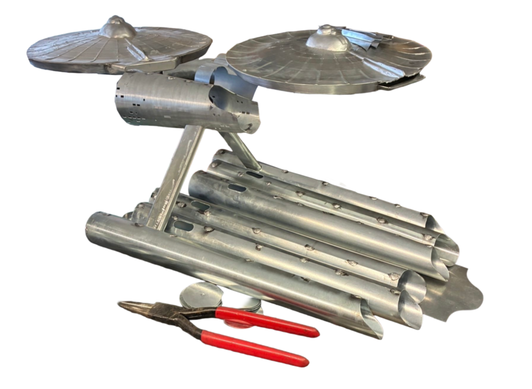Ein silbernes Modell eines Raumschiffs, das der Star Trek Enterprise ähnelt und aus Metallteilen und Rohren besteht, ist neben einer Zange mit rotem Griff auf weißem Hintergrund ausgestellt.