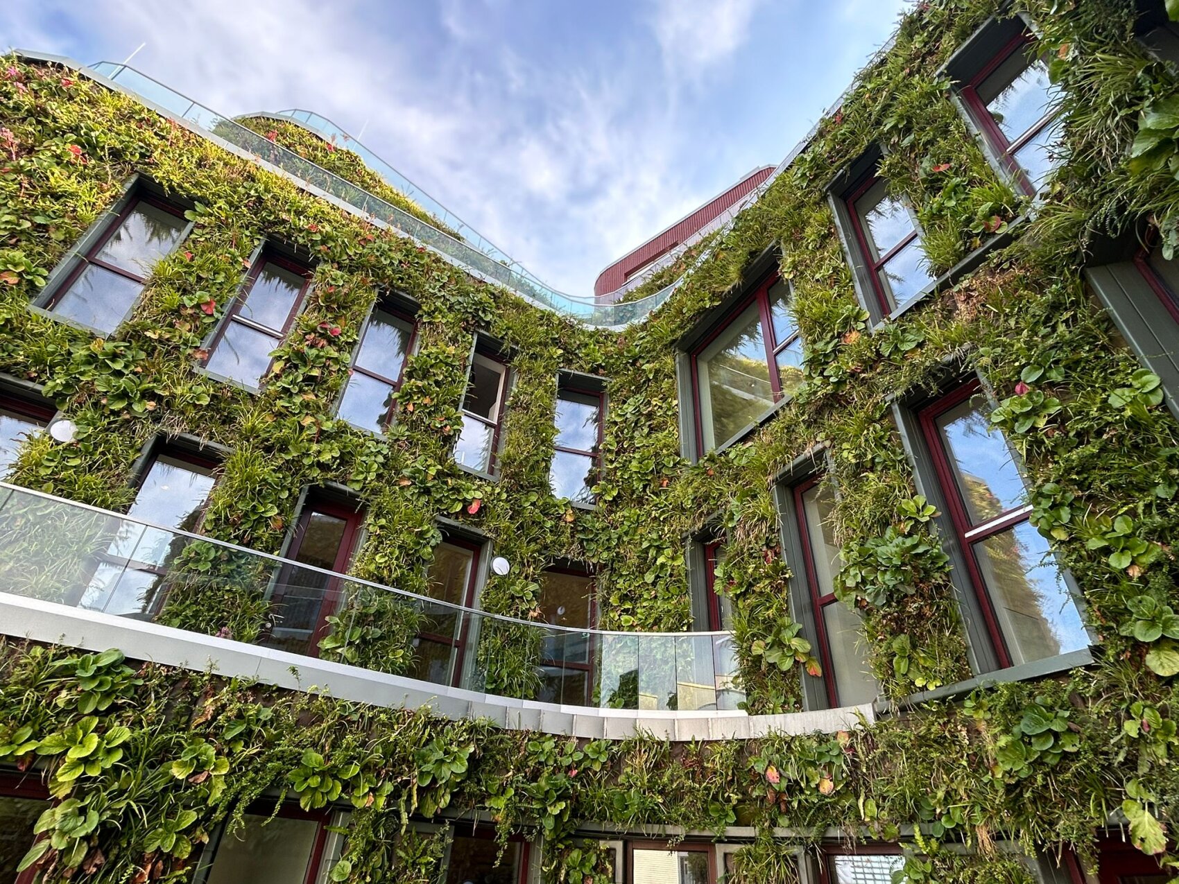 Ein mehrstöckiges Gebäude, dessen Wände mit üppigen grünen Pflanzen bedeckt sind, mit großen Fenstern und Balkonen, mit Blick auf einen Innenhof unter einem blauen Himmel mit einigen Wolken.